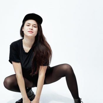 Young stylish dark-haired girl dressed in a black top, shorts, tights and cap  sits in a squat  on the white background in the studio .